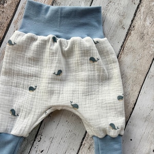 Könnte beinhalten: Babyhose mit einem weißen Stoff, der mit kleinen blauen Walillustrationen bedeckt ist. Die Hose hat einen hellblauen Bund und Bündchen. Der Stoff hat ein strukturiertes, geknittertes Aussehen.
