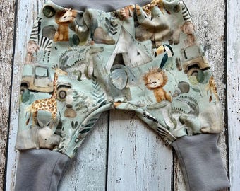 May include: Baby pants with a safari animal print in shades of green, tan, and gray. The print features lions, giraffes, zebras, and other animals, along with safari vehicles and tents. The pants have gray cuffs and a gray waistband.