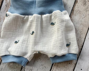 May include: White baby shorts with a blue waistband and cuffs. The shorts are decorated with small blue whale designs. The fabric appears to be a soft, textured material, suitable for infants and toddlers.