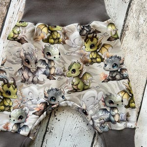 Puede incluir: Pantalones de bebé con cintura y puños grises. Los pantalones presentan un fondo beige claro con un estampado repetido de dragones de dibujos animados en varios colores, incluyendo verde, gris y amarillo. Los dragones tienen ojos grandes y alas.