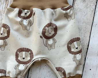 Puede incluir: Pantalones de bebé con cintura y puños color canela. La tela blanquecina presenta un estampado repetido de leones de dibujos animados en marrón y beige. La tela tiene un aspecto moteado.