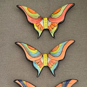 May include: Three ceramic butterfly wall decorations in a vertical row. The top butterfly is orange, yellow, and green. The middle butterfly is blue, yellow, and orange. The bottom butterfly is blue, orange, and yellow.