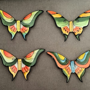 Puede incluir: Cuatro decoraciones de pared de mariposa de cerámica con diseños pintados coloridos. Las mariposas son en tonos de verde, amarillo, naranja y azul.