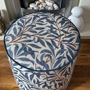 May include: A circular ottoman upholstered in fabric with a blue and cream leaf design. The pattern shows blue leaves and brown branches on a cream backdrop. It has a dark blue trim.