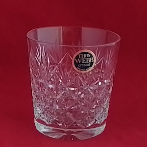 Puede incluir: Un vaso de cristal transparente con un diseño de estrella. El vaso tiene una base gruesa y un borde ligeramente ensanchado. Una pequeña etiqueta redonda con el texto "THOS WEBB crystal BRITISH MADE" es visible.