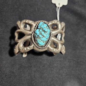 May include: A silver cuff bracelet featuring a central, rough-cut turquoise stone with dark matrix veining. The stone is encased in a twisted rope border, surrounded by an openwork metal design with curved, scroll-like patterns and a white price tag.