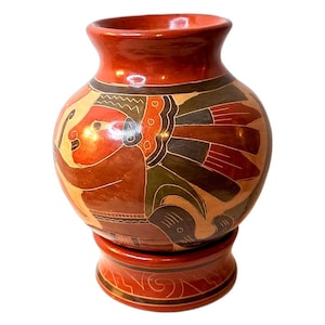 Puede incluir: Un jarrón de terracota pintado a mano con una base elevada. El jarrón presenta una figura estilizada con un tocado y detalles intrincados en tonos rojos, marrones, verdes y negros.