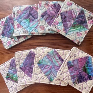 May include: Eight handmade quilted coasters featuring a floral design in shades of purple, pink, and green. The coasters are made from a cream-coloured fabric with a subtle floral pattern.