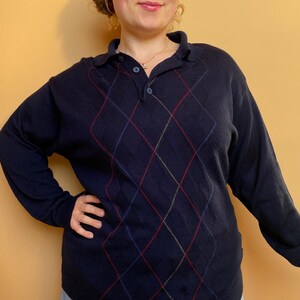 Könnte beinhalten: Ein marineblauer Pullover mit einem Rautenmuster in roter, blauer und goldener Farbe. Der Pullover hat einen Kragen und einen Knopfverschluss.