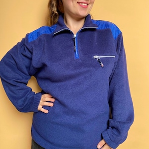 Op de afbeelding: Een blauwe fleece trui met een halve ritssluiting en een blauw nylon paneel op de schouders. De trui heeft een ritszakje aan de linkerkant.