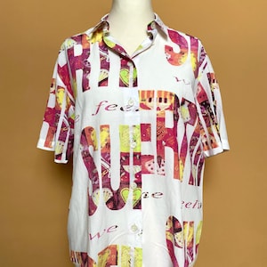 Op de afbeelding: Wit overhemd met knopen en een kleurrijke print van de woorden "feel" en "super" in roze, geel en groen.