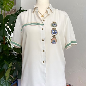 Könnte beinhalten: Ein weißes Hemd mit Knöpfen, kurzen Ärmeln und einem grün-rosa Streifen über der Brust. Das Hemd hat eine Reihe von dekorativen Knöpfen und eine goldene Kettenkette mit einem Anhänger.