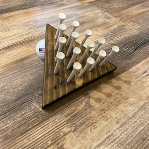 May include: A wooden triangular game board with 15 white golf tees arranged in a pyramid shape. A white golf ball is in the foreground.