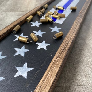 May include: A wooden framed sign with a black background and white stars, a blue and white thin blue line, and bullet casings used as push pins.