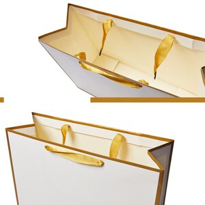 May include: White gift bag with gold trim and gold ribbon handles. The bag is open and the inside is lined with a cream colored paper.
