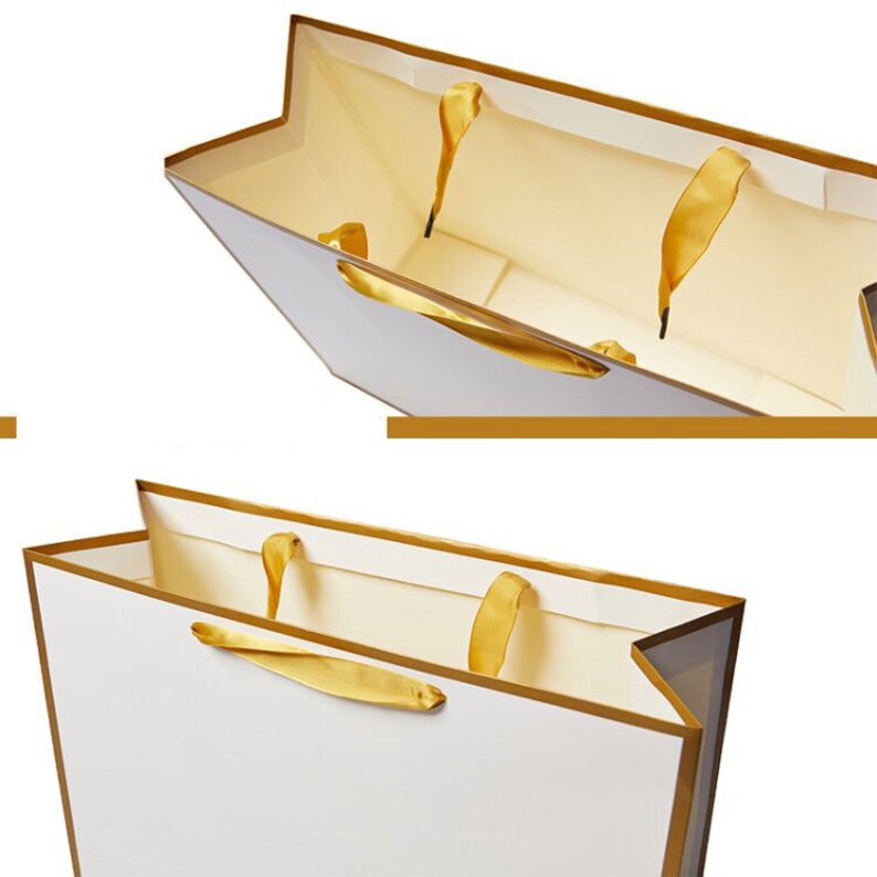 May include: White gift bag with gold trim and gold ribbon handles. The bag is open and the inside is lined with a cream colored paper.