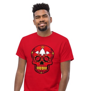 May include: A red t-shirt with a sugar skull design featuring a white arrowhead and black details. The skull is surrounded by a floral pattern.