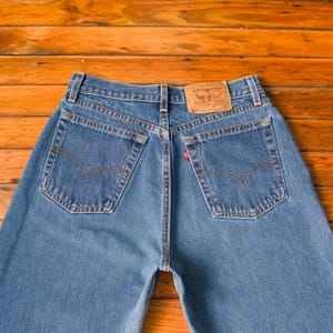 Könnte beinhalten: Blaue Jeans aus Denim mit klassischem Fünf-Taschen-Design. Merkmale sind ein rotes Levi's-Etikett auf der Gesäßtasche und ein brauner Lederaufnäher. Die Jeans werden auf einer Holzoberfläche präsentiert.
