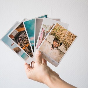 May include: A hand holds a stack of five colorful photographs. The top photo shows a person carrying a large bundle of dried grass on their head. The photo below it shows a group of people walking down a street. The other photos show a variety of landscapes and scenes.