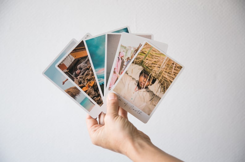 May include: A hand holds a stack of five colorful photographs. The top photo shows a person carrying a large bundle of dried grass on their head. The photo below it shows a group of people walking down a street. The other photos show a variety of landscapes and scenes.
