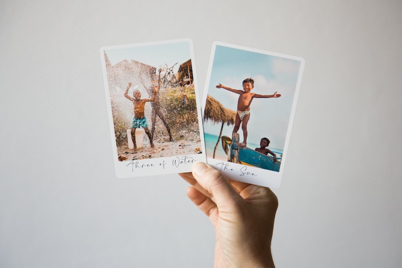 May include: Two Polaroid-style photos with handwritten text. The first photo shows two children playing in a water spray. The text below the photo reads "Three of Water". The second photo shows a child standing on a boat with arms outstretched. The text below the photo reads "The Sun".