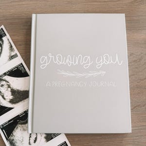 May include: A light gray hardcover journal with white text that reads growing you A PREGNANCY JOURNAL and a small leaf graphic. Next to the journal are two black and white ultrasound scans showing fetal development images on a wooden surface.