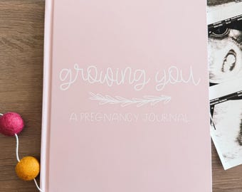 May include: A light pink hardcover journal with the white text "growing you" and "A PREGNANCY JOURNAL" printed on the front cover, accompanied by a white line drawing of a leafy branch. A string of small, colorful felt balls sits nearby.