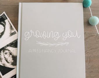 Könnte beinhalten: Ein hellgraues Schwangerschaftstagebuch mit festem Einband, auf dessen Vorderseite der weiße Schriftzug "growing you" und "A PREGNANCY JOURNAL" sowie eine weiße Strichzeichnung eines Zweiges und mehrere schwarz-weiße Ultraschallbilder zu sehen sind.