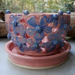 May include: A pink ceramic planter with a matching saucer. The planter features a floral design with blue flowers and small blue accents around the rim. The planter is designed for indoor or outdoor use.