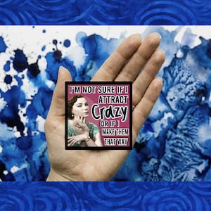 May include: A square black sticker with the text "I'M NOT SURE IF I ATTRACT Crazy OR IF I MAKE THEM THAT WAY" and a vintage image of a woman. The sticker is held in a hand against a blue and white watercolor background.