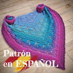 May include: A handmade crochet shawl in a gradient of blue, turquoise, and purple hues. The shawl features intricate openwork patterns and is adorned with two crocheted rose accents. The text "Patrón en ESPAÑOL" is visible.