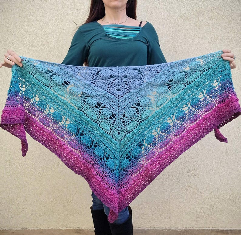 Pu&ograve; includere: Uno scialle triangolare all'uncinetto con un gradiente di rosa, blu e viola. Lo scialle presenta un delicato motivo a pizzo e un bordo a frangia.
