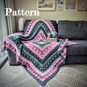May include: A crocheted blanket in pink, teal, and grey, draped over a grey sofa. The blanket features a detailed, geometric pattern with scalloped edges. The word "Pattern" is in the upper left corner.