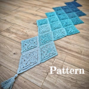 Puede incluir: Camino de mesa de ganchillo azul hecho a mano, con un patrón de diamantes y una borla. El camino de mesa presenta un degradado de tonos azules, de claro a oscuro, y se muestra sobre una superficie de madera. La palabra "Pattern" está escrita en la imagen.