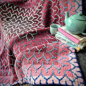 Puede incluir: Una manta de ganchillo hecha a mano en tonos burdeos, rosa y azul. La manta presenta un detallado patrón floral en el centro y un diseño geométrico repetitivo en los bordes. Una tetera, una taza y libros de color turquesa están sobre la manta.