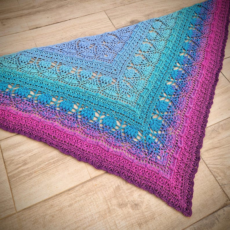 Pu&ograve; includere: Uno scialle triangolare all'uncinetto con un gradiente di colori blu, turchese e rosa. Lo scialle presenta un delicato motivo a pizzo e un bordo a festoni.