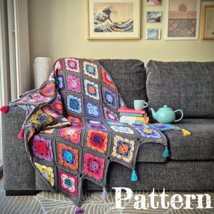 Könnte beinhalten: Eine handgefertigte Häkeldecke mit einem farbenfrohen quadratischen Muster, drapiert über einem grauen Sofa. Die Decke hat Quadrate in verschiedenen Farben, darunter Rosa, Blau, Orange und Lila, mit grauen Rändern. Die Decke hat Quasten an den Ecken. Das Wort "Pattern" ist sichtbar.