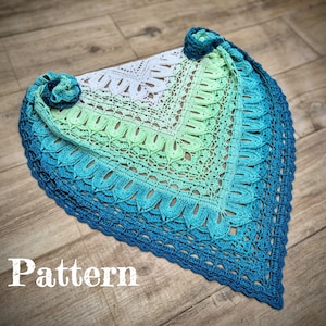 Può includere: Uno scialle all'uncinetto in sfumature di blu e verde, con un delicato motivo traforato. Lo scialle presenta un bordo smerlato e due decorazioni floreali. La parola "Pattern" è visualizzata in testo bianco.