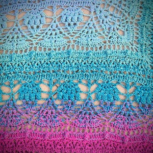 Pu&ograve; includere: Primo piano di una coperta all'uncinetto con un gradiente di colori blu e rosa. La coperta presenta un delicato motivo a pizzo con un design floreale ripetitivo.