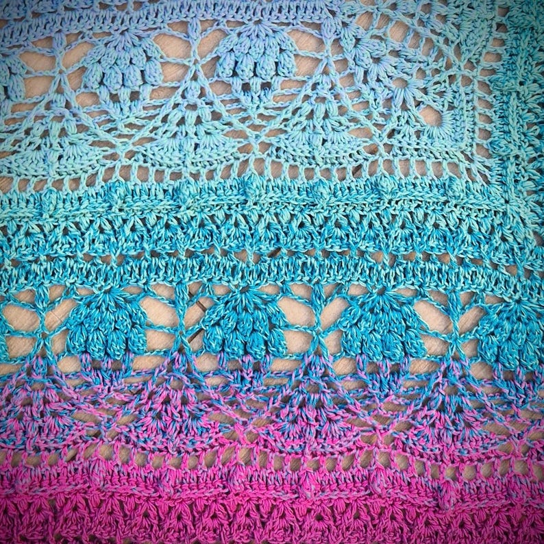 Pu&ograve; includere: Primo piano di una coperta all'uncinetto con un gradiente di colori blu e rosa. La coperta presenta un delicato motivo a pizzo con un design floreale ripetitivo.