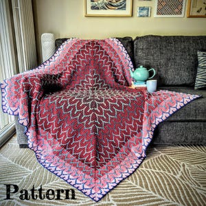 Könnte beinhalten: Eine handgefertigte Decke mit einem sich wiederholenden Blattmuster in Rot, Rosa und Blau, drapiert über einem grauen Sofa. Die Decke hat einen dunkelblauen Rand. Eine türkisfarbene Teekanne und eine Tasse stehen auf dem Sofa.