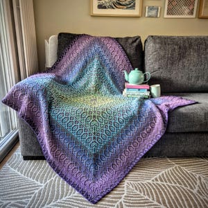 Può includere: Una coperta all'uncinetto quadrata caratterizzata da una sfumatura di colore che va dal giallo centrale al turchese, blu e viola intenso. La coperta presenta un complesso motivo geometrico ripetitivo su tutta la superficie.