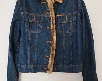 Peut inclure: Veste en jean bleue avec col, bordure et poignets en fausse fourrure marron. La veste a deux poches poitrine et des boutons. La veste est suspendue à un cintre gris sur fond blanc.