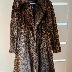 Könnte beinhalten: Ein doppelreihiger Mantel mit Leopardenmuster und breitem Kragen. Der Mantel ist braun und schwarz mit einer Kunstfellstruktur. Der Mantel hängt an einem Kleiderbügel. Der Mantel hat einen Knopfverschluss.