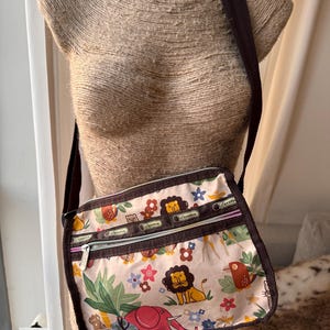May include: A white and brown crossbody bag with a jungle animal print. The bag has three zippered compartments and a brown adjustable strap. The bag features a variety of animals including lions, elephants, birds, and monkeys.