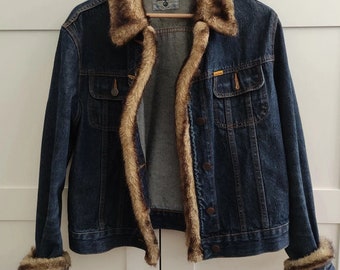 Peut inclure: Veste en jean bleu foncé avec col, poignets et doublure en fausse fourrure marron. La veste a deux poches avant et une fermeture boutonnée. La veste est suspendue à un cintre en bois.