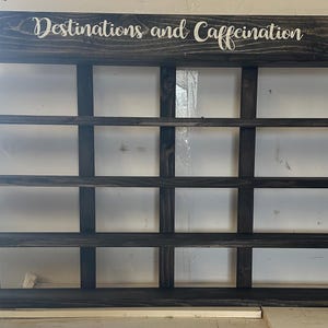 May include: A dark stained wooden display shelf with four horizontal rows and four vertical dividers creating sixteen rectangular compartments. The top header features the white text "Destinations and Caffeination" in a cursive script font.