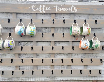 May include: A gray wooden coffee mug rack with black metal hooks. The rack has the words "Coffee Travels" written on it in white paint. There are several colorful coffee mugs hanging on the hooks.