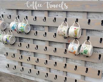 May include: A gray wooden coffee mug rack with black metal hooks. The rack has the words "Coffee Travels" written on the top. There are several colorful mugs hanging on the hooks.