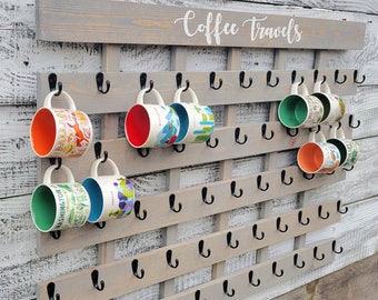 May include: A wooden coffee mug rack with the words "Coffee Travels" painted on the top. The rack has multiple hooks for hanging mugs. There are several colorful mugs hanging on the hooks.
